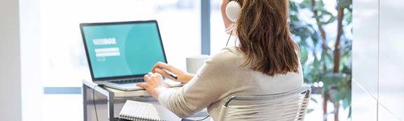 A woman sitting viewing a laptop and wearing earphones.